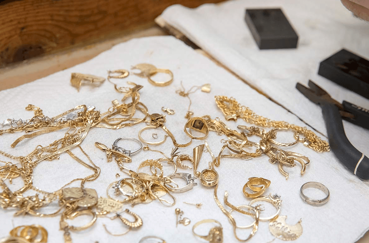 Ungeordnete Haufen aus Gold- und Silberschmuck auf weißem Tuch: Ketten, Ringe, Anhänger, Ohrringe, Werkzeugzangen im Hintergrund